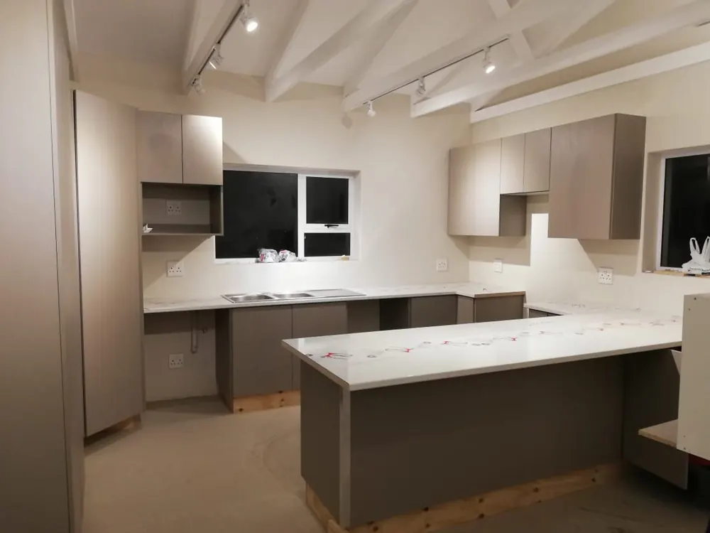 A new kitchen mid-installation, taupe doors and a white stone worktop still under protective film, beneath white roof trusses.
