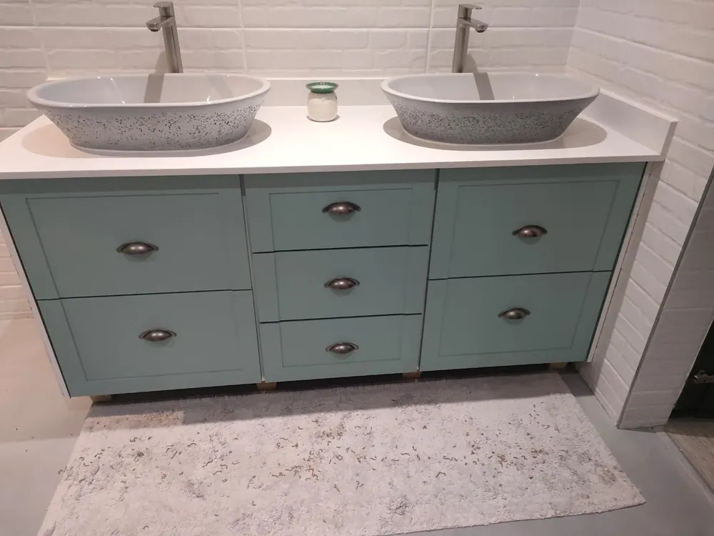 A double bathroom vanity in sage green shaker doors with cup handles, topped in white stone with two speckled basins.